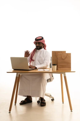 A business meeting held via laptop, the concept of electronic marketing and online orders, Saudi professions and jobs, a portrait of an Arab Gulf Saudi man wearing traditional attire and a ghutrah using a laptop with shopping bags in front of him, browsing social media, using modern technological devices, remote work, white background.