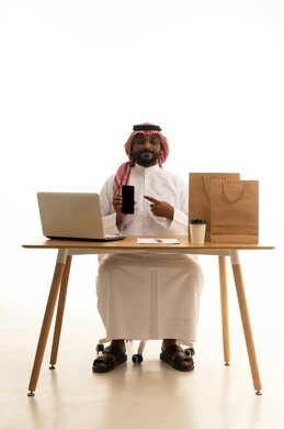 A portrait of a Saudi Arabian Gulf man wearing traditional attire and a shemagh, using a laptop while pointing to a mobile phone in front of him, with shopping bags, a display of a blank black screen, browsing social media, using modern technology devices, working remotely, the concept of electronic marketing and online orders, with a white background.