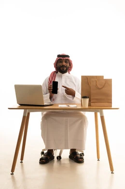 إستخدام اجهزة تقنية حديثة ، العمل عن بعد ، عرض شاشة سوداء فارغة، تصفح وسائل التواصل الاجتماعي،مفهوم التسويق الإلكتروني وطلبات الاونلاين ،بورتريه لرجل عربي خليجي سعودي يرتدي الزى التقليدي والشماغ يستخدم جهاز الحاسوب المحمول يشير الى الهاتف المحمول امامه اكياس التسوق الورقية ،خلفية بيضاء