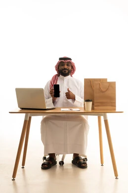تصفح وسائل التواصل الاجتماعي، عرض شاشة سوداء فارغة،إستخدام اجهزة تقنية حديثة ، العمل عن بعد ، مفهوم التسويق الإلكتروني وطلبات الاونلاين ،بورتريه لرجل عربي خليجي سعودي يرتدي الزى التقليدي والشماغ يستخدم جهاز الحاسوب المحمول يشير الى الهاتف المحمول امامه اكياس التسوق الورقية ،خلفية بيضاء