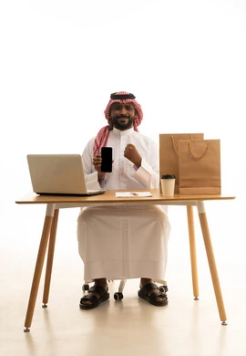 عرض شاشة سوداء فارغة، تصفح وسائل التواصل الاجتماعي، إستخدام اجهزة تقنية حديثة ، العمل عن بعد ، مفهوم التسويق الإلكتروني وطلبات الاونلاين ،بورتريه لرجل عربي خليجي سعودي يرتدي الزى التقليدي والشماغ يستخدم جهاز الحاسوب المحمول يشير الى الهاتف المحمول امامه اكياس التسوق الورقية ،خلفية بيضاء