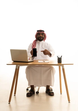 بورتريه لرجل عربي خليجي سعودي يرتدي الزى التقليدي والشماغ يستخدم جهاز الحاسوب المحمول ، اجراء مكالمة هاتفية، إستخدام اجهزة تقنية حديثة ، العمل عن بعد ،عرض شاشة سوداء فارغة عبر الجوال، عقد إجتماع عمل عبر اللابتوب ،تصفح وسائل التواصل الاجتماعي، خلفية بيضاء
