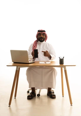 Remote work, making a phone call, using modern technological devices, displaying a blank black screen on the mobile, holding a business meeting via laptop, a portrait of a Saudi Arabian Gulf man wearing traditional attire and a ghutrah using a laptop, browsing social media, white background.