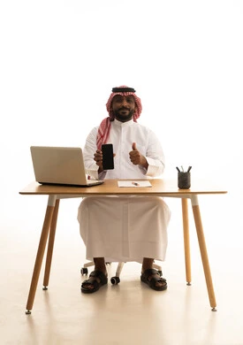 إستخدام اجهزة تقنية حديثة ، العمل عن بعد ،عرض شاشة سوداء فارغة عبر الجوال، عقد إجتماع عمل عبر اللابتوب ، بورتريه لرجل عربي خليجي سعودي يرتدي الزى التقليدي والشماغ يستخدم جهاز الحاسوب المحمول ، تصفح وسائل التواصل الاجتماعي، خلفية بيضاء
