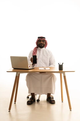 عرض شاشة سوداء فارغة عبر الجوال، عقد إجتماع عمل عبر اللابتوب ، اجراء مكالمة هاتفية، إستخدام اجهزة تقنية حديثة ، العمل عن بعد ،بورتريه لرجل عربي خليجي سعودي يرتدي الزى التقليدي والشماغ يستخدم جهاز الحاسوب المحمول ، تصفح وسائل التواصل الاجتماعي، خلفية بيضاء