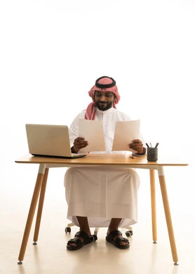 مهن ووظائف مكتبيه، مفهوم ادارة الاعمال عن بعد، عقد إجتماع عمل عبر اللابتوب ،  بورتريه لرجل عربي خليجي سعودي يرتدي الزى التقليدي والشماغ يحمل بيده الاوراق ويقوم بتدقيق العمل امام جهاز الحاسوب المحمول, خلفية بيضاء