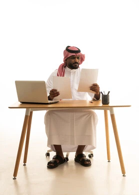 دمج العمل مع التقنية والتكنولوجيا ، مفهوم ادارة الاعمال عن بعد، مهن ووظائف مكتبيه،  بورتريه لرجل عربي خليجي سعودي يرتدي الزى التقليدي والشماغ يحمل بيده الاوراق ويقوم بتدقيق العمل امام جهاز الحاسوب المحمول, خلفية بيضاء