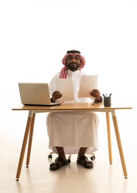 A portrait of a Saudi Arabian Gulf man wearing traditional attire and a shemagh, holding papers in his hand and reviewing work in front of a laptop, office professions and jobs, holding a business meeting via the laptop, the concept of remote business management, white background.