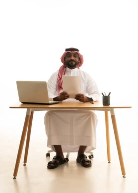 استخدام جهاز تقني حديث ،مفهوم ادارة الاعمال عن بعد، عقد إجتماع عمل عبر اللابتوب ، مهن ووظائف مكتبيه،  بورتريه لرجل عربي خليجي سعودي يرتدي الزى التقليدي والشماغ يحمل بيده الاوراق ويقوم بتدقيق العمل امام جهاز الحاسوب المحمول, خلفية بيضاء