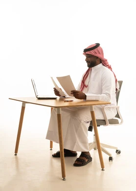 Using white papers in work planning, holding a business meeting via laptop, the concept of remote business management, office professions and jobs, a portrait of a Saudi Arabian Gulf man wearing traditional attire and a ghutrah holding papers in his hand and reviewing work in front of a laptop, white background.