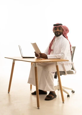 The concept of remote business management, holding a business meeting via laptop, office professions and jobs, a portrait of a Saudi Arabian Gulf man wearing traditional attire and a ghutrah, holding papers in his hand and reviewing work in front of a laptop, white background.