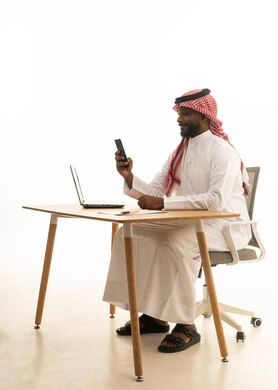 Browsing social media, using modern technology devices, working remotely, displaying a blank black screen on the mobile, holding a business meeting via laptop, a portrait of a Saudi Arabian Gulf man wearing traditional attire and a ghutrah using a laptop, white background.