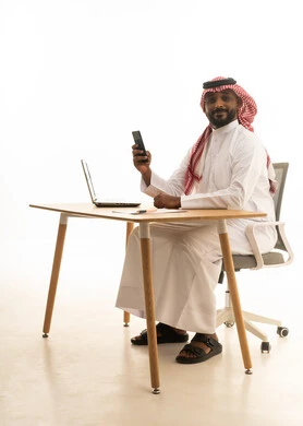Using modern technology devices, remote work, displaying a blank black screen on the mobile phone, holding a business meeting via laptop, a portrait of a Saudi Arabian Gulf man wearing traditional attire and a ghutrah using a laptop, browsing social media, white background.