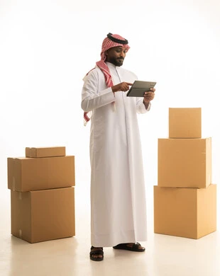 A portrait of a Saudi Arabian Gulf man wearing traditional attire and a ghutrah, carrying boxes of household items while holding a tablet in his hand. It represents owning a new home and family stability, modern housing and ownership, using modern technological devices, organizing and arranging the home, a full-body image, with a white background.