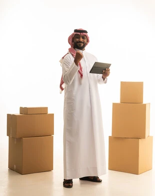 رفع قبضة اليد بتعابير الانتصار،استخدام اجهزه تقنية حديثة ، ترتيب وتنظيم المنزل، بورتريه لرجل عربي خليجي سعودي يرتدي الزى التقليدي والشماغ  يقوم بنقل صناديق الأغراض المنزلية يمسك بيده جهاز التابلت ، امتلاك سكن جديد والاستقرار العائلي، اسكان وتمليك حديث ، صورة الجسم بالطول الكامل، خلفية بيضاء