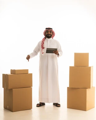 Modern housing and ownership, using modern technological devices, arranging and organizing the home, a portrait of a Saudi Arabian Gulf man wearing traditional attire and a ghutrah, carrying boxes of household items while holding a tablet in his hand, owning a new home and family stability, full-body image, white background.