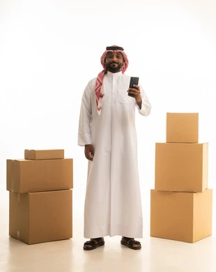 Modern housing and ownership, using a mobile phone, making a phone call, arranging and organizing the home, a portrait of a Saudi Arabian Gulf man wearing traditional attire and a ghutrah, carrying boxes of household items while holding a mobile device, owning a new home and family stability, full-body image, white background.