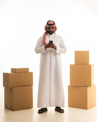 Using a mobile phone, making a phone call, arranging and organizing the house, a portrait of a Saudi Arabian Gulf man wearing traditional attire and a ghutrah, carrying boxes of household items while holding a mobile device, owning a new home and family stability, modern housing and ownership, full-body image, white background.