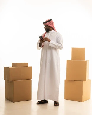 Browsing social media, using a mobile phone, organizing and arranging the home, a portrait of a Saudi Arabian Gulf man wearing traditional attire and a ghutrah, carrying boxes of household items while holding a mobile device, owning a new home and family stability, modern housing and ownership, full-body image, white background.