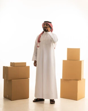 اجراء مكالمه هاتفيه،  ترتيب وتنظيم المنزل، بورتريه لرجل عربي خليجي سعودي يرتدي الزى التقليدي والشماغ  يقوم بنقل صناديق الأغراض المنزلية يمسك بيده جهاز الجوال ، امتلاك سكن جديد والاستقرار العائلي، اسكان وتمليك حديث ، صورة الجسم بالطول الكامل، خلفية بيضاء