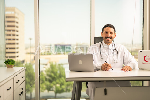 استخدام حقيبة الاسعافات الاوليه و جهاز الحاسوب في الطب، متابعة حالات المرضى عبر الاجهزة و التقنيات الحديثة,  طبيب عربي خليجي سعودي مبتسم يرتدي المعطف الأبيض يقوم بكتابة وتدوين التقارير والحالات الصحيه
