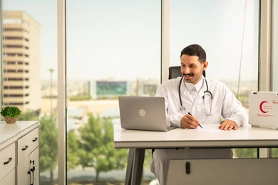 Using a first aid kit and a computer in medicine, following up on patients' cases using modern devices and technologies, a smiling Saudi Arabian Gulf doctor wearing a white coat writing and recording reports and health cases