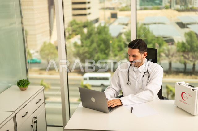 Using a first aid kit and a computer in medicine, following up on patients' cases using modern devices and technologies, a smiling Saudi Arabian Gulf doctor wearing a white coat writing and recording reports and health cases