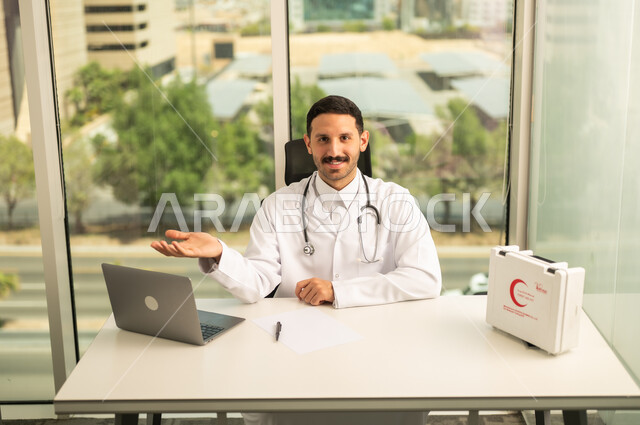 متابعة حالات المرضى عبر الاجهزة و التقنيات الحديثة, استخدام حقيبة الاسعافات الاوليه و جهاز الحاسوب في الطب، طبيب عربي خليجي سعودي مبتسم يرتدي المعطف الأبيض ينظر إلى الكاميرا بإيماءات الثقة بالنفس، تسجيل التقارير والحالات الصحيه