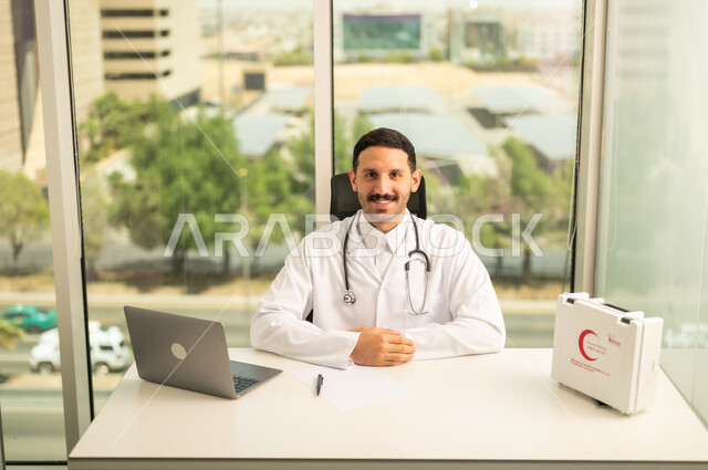 متابعة حالات المرضى عبر الاجهزة و التقنيات الحديثة, استخدام حقيبة الاسعافات الاوليه و جهاز الحاسوب في الطب، طبيب عربي خليجي سعودي مبتسم يرتدي المعطف الأبيض ينظر إلى الكاميرا بإيماءات الثقة بالنفس، تسجيل التقارير والحالات الصحيه