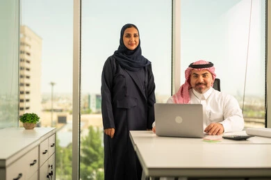 الوحدة والعمل الجماعي، استخدام جهاز تقني حديث، موظفة تقوم بتقديم المشاريع للمدير لتدقيقها عبر جهاز اللابتوب، تطوير ودراسة وتحليل المشاريع الجديدة، موظف عربي خليجي سعودي يرتدي الثوب والشماغ التقليدي يجلس خلف المكتب داخل مقر الشركة، مهنة ووظيفة سعودية مكتبية