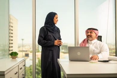 استخدام جهاز تقني حديث، موظفة تقوم بتقديم المشاريع للمدير لتدقيقها عبر جهاز اللابتوب، تطوير ودراسة وتحليل المشاريع الجديدة، موظف عربي خليجي سعودي يرتدي الثوب والشماغ التقليدي يجلس خلف المكتب داخل مقر الشركة، الوحدة والعمل الجماعي، مهنة ووظيفة سعودية مكتبية