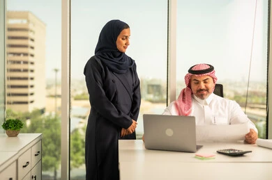 الوحدة والعمل الجماعي، استخدام جهاز تقني حديث، موظفة تقوم بتقديم المشاريع للمدير لتدقيقها عبر جهاز اللابتوب، تطوير ودراسة وتحليل المشاريع الجديدة، موظف عربي خليجي سعودي يرتدي الثوب والشماغ التقليدي يجلس خلف المكتب داخل مقر الشركة، مهنة ووظيفة سعودية مكتبية