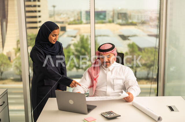 استخدام جهاز تقني حديث، موظفة تقوم بتقديم المشاريع للمدير لتدقيقها عبر جهاز اللابتوب، تطوير ودراسة وتحليل المشاريع الجديدة، موظف عربي خليجي سعودي يرتدي الثوب والشماغ التقليدي يجلس خلف المكتب داخل مقر الشركة، الوحدة والعمل الجماعي، مهنة ووظيفة سعودية مكتبية