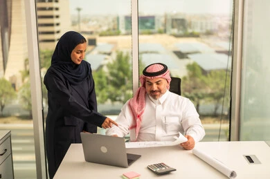 استخدام جهاز تقني حديث، موظفة تقوم بتقديم المشاريع للمدير لتدقيقها عبر جهاز اللابتوب، تطوير ودراسة وتحليل المشاريع الجديدة، موظف عربي خليجي سعودي يرتدي الثوب والشماغ التقليدي يجلس خلف المكتب داخل مقر الشركة، الوحدة والعمل الجماعي، مهنة ووظيفة سعودية مكتبية