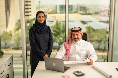 Unity and teamwork, using modern technology, an employee presenting projects to the manager for review via laptop, developing, studying and analyzing new projects, a Saudi Gulf Arab employee wearing the traditional thobe and shemagh sitting behind a desk inside the company headquarters, a Saudi office profession and job