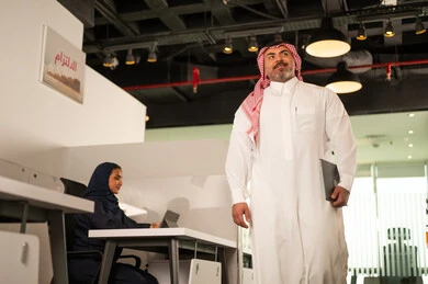 مهنة ووظيفة سعودية مكتبية، استخدام جهاز تقني حديث، تطوير ودراسة وتحليل المشاريع الجديدة، موظفة عربية خليجية سعودية محجبة تجلس خلف المكتب تقوم باستخدام الحاسوب المحمول، موظف عربي خليجي سعودي يرتدي الثوب والشماغ في مقر الشركة