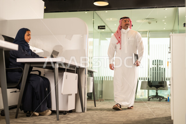 مهنة ووظيفة سعودية مكتبية، استخدام جهاز تقني حديث، تطوير ودراسة وتحليل المشاريع الجديدة، موظفة عربية خليجية سعودية محجبة تجلس خلف المكتب تقوم باستخدام الحاسوب المحمول، موظف عربي خليجي سعودي يرتدي الثوب والشماغ في مقر الشركة