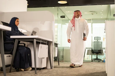 مهنة ووظيفة سعودية مكتبية، استخدام جهاز تقني حديث، تطوير ودراسة وتحليل المشاريع الجديدة، موظفة عربية خليجية سعودية محجبة تجلس خلف المكتب تقوم باستخدام الحاسوب المحمول، موظف عربي خليجي سعودي يرتدي الثوب والشماغ في مقر الشركة