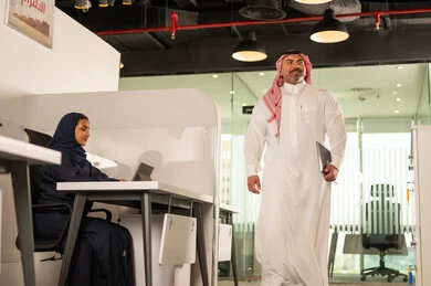 Saudi office profession and job, using modern technical equipment, developing, studying and analyzing new projects, a veiled Saudi Arabian Gulf female employee sitting behind a desk using a laptop, a Saudi Arabian Gulf male employee wearing a thobe and shemagh at the company headquarters