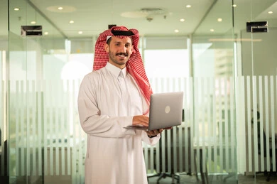 استخدام جهاز الحاسوب المحمول لإنجاز المهام باحترافية، التوظيف في المكاتب والشركات السعودية، توفير بيئة عمل مريحة ومناسبة، النظر إلى الكاميرا بإيماءات السرور، رجل عربي خليجي سعودي يرتدي الثوب والشماغ التقليدي يقوم بالعمل عبر اللابتوب داخل مقر الشركة