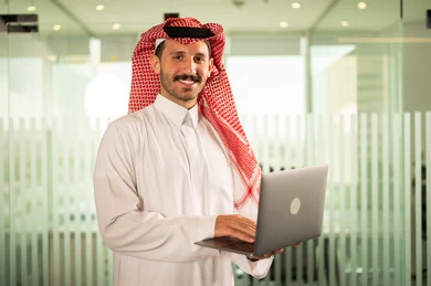 التوظيف في المكاتب والشركات السعودية، توفير بيئة عمل مريحة ومناسبة، النظر إلى الكاميرا بإيماءات السرور، استخدام جهاز الحاسوب المحمول لإنجاز المهام باحترافية، رجل عربي خليجي سعودي يرتدي الثوب والشماغ التقليدي يقوم بالعمل عبر اللابتوب داخل مقر الشركة