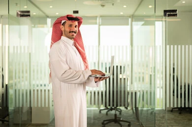 استخدام جهاز الحاسوب المحمول لإنجاز المهام باحترافية، التوظيف في المكاتب والشركات السعودية، توفير بيئة عمل مريحة ومناسبة، النظر إلى الكاميرا بإيماءات السرور، رجل عربي خليجي سعودي يرتدي الثوب والشماغ التقليدي يقوم بالعمل عبر اللابتوب داخل مقر الشركة