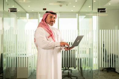 Recruitment in Saudi offices and companies, providing a comfortable and suitable work environment, looking at the camera with gestures of pleasure, using a laptop to complete tasks professionally, a Saudi Arabian Gulf man wearing the traditional thobe and shemagh working on a laptop inside the company headquarters