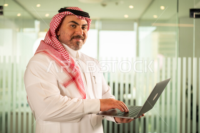 Recruitment in Saudi offices and companies, providing a comfortable and suitable work environment, looking at the camera with gestures of pleasure, using a laptop to complete tasks professionally, a Saudi Arabian Gulf man wearing the traditional thobe and shemagh working on a laptop inside the company headquarters