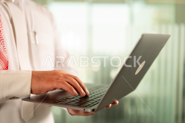 توفير بيئة عمل مريحة ومناسبة، استخدام جهاز الحاسوب المحمول لإنجاز المهام باحترافية، التوظيف في المكاتب والشركات السعودية، صورة مقربة لرجل عربي خليجي سعودي يرتدي الثوب والشماغ التقليدي يقوم بالعمل عبر اللابتوب داخل مقر الشركة