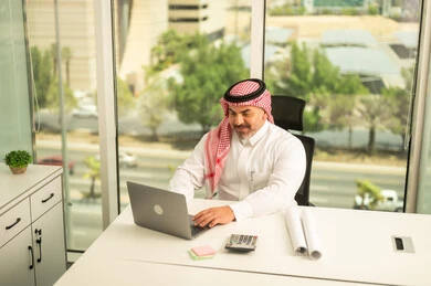 Office jobs and professions, gestures of integration and focus, providing a comfortable and suitable work environment, using a laptop to complete tasks professionally, a Saudi Arabian Gulf man wearing a traditional thobe and shemagh sitting in an office working on a laptop, employment in Saudi offices and companies