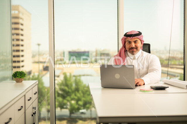 مهن وأعمال مكتبية، إيماءات الاندماج والتركيز، توفير بيئة عمل مريحة ومناسبة، استخدام جهاز الحاسوب المحمول لإنجاز المهام باحترافية، رجل عربي خليجي سعودي يرتدي الثوب والشماغ التقليدي يجلس في المكتب يقوم بالعمل عبر اللابتوب، التوظيف في المكاتب والشركات السعودية