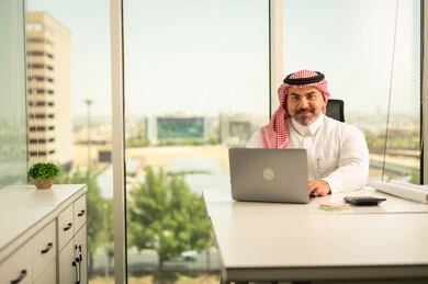 مهن وأعمال مكتبية، إيماءات الاندماج والتركيز، توفير بيئة عمل مريحة ومناسبة، استخدام جهاز الحاسوب المحمول لإنجاز المهام باحترافية، رجل عربي خليجي سعودي يرتدي الثوب والشماغ التقليدي يجلس في المكتب يقوم بالعمل عبر اللابتوب، التوظيف في المكاتب والشركات السعودية