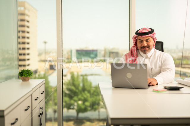 Office jobs and professions, gestures of integration and focus, providing a comfortable and suitable work environment, using a laptop to complete tasks professionally, a Saudi Arabian Gulf man wearing a traditional thobe and shemagh sitting in an office working on a laptop, employment in Saudi offices and companies