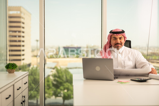 تدقيق مخططات المشاريع، إيماءات الاندماج والتركيز، توفير بيئة عمل مريحة ومناسبة، استخدام جهاز الحاسوب المحمول لإنجاز المهام باحترافية، رجل عربي خليجي سعودي يرتدي الثوب والشماغ التقليدي يجلس في المكتب يقوم بالعمل عبر اللابتوب، التوظيف في المكاتب والشركات السعودية